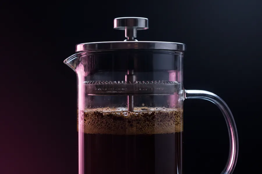 Prensa francesa de vidrio llena de café oscuro, con el émbolo arriba y la costra de posos flotando.