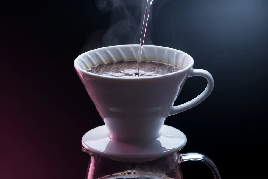 Cono V60 blanco sobre jarra de vidrio, con un hilo de agua cayendo sobre el lecho de café.